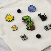 A set of colorful enamel pins displayed on a cream-colored canvas tote bag. The pins feature whimsical designs including a yellow smiling sun, a green Venus Flytrap plant in a pot, a black cat, a yellow and brown stack of pancakes, a purple and blue spiral, a retro game controller, and small black dome-shaped charms.