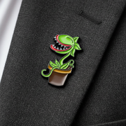 A small enamel pin shaped like a green Venus flytrap plant in a brown pot, pinned onto a dark charcoal gray suit jacket lapel next to a white shirt cuff.